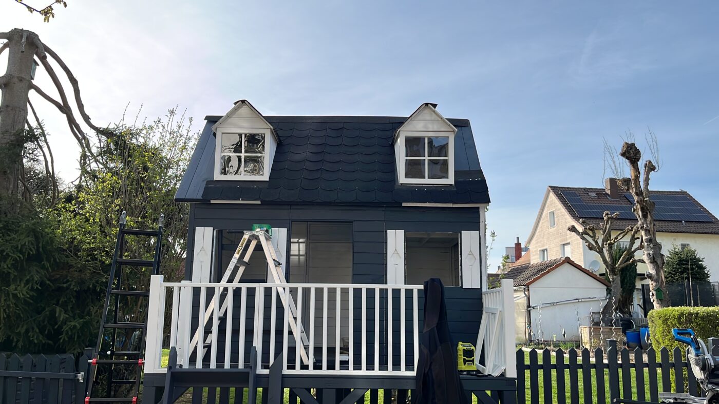 Ein teilweise aufgebautes, dunkel gestrichenes Spielhaus mit weißem Balkon und zwei kleinen Gaubenfenstern. Eine Leiter lehnt am Geländer, Bauwerkzeuge stehen bereit. Im Hintergrund sind Wohnhäuser und ein frisch errichteter Gartenzaun zu sehen.