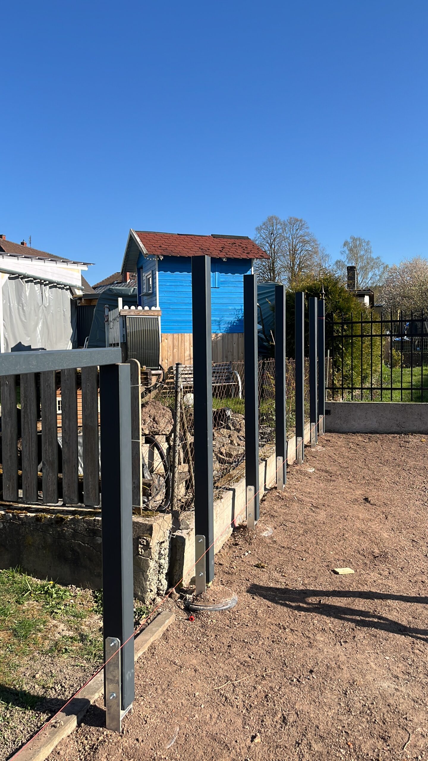 Blick auf eine Baustelle mit frisch gesetzten Metallpfosten für einen Gartenzaun aus Holz. Im Hintergrund stehen ein blau gestrichenes Spielhaus und mehrere Gartenhäuser. Eine gespannte Richtschnur verläuft am Boden entlang der Zaunlinie, die sich an ein Gefälle im Gelände anpasst.