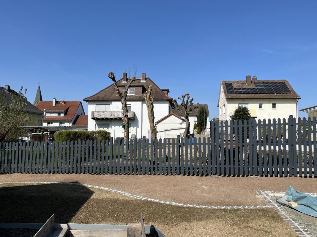 Ein dunkel lasierter Gartenzaun aus Holz zieht sich entlang eines Grundstücks vor mehreren Wohnhäusern. Der Zaun ist auf der rechten Seite höher, um Sichtschutz zu bieten. Im Vordergrund sieht man einen hell gepflasterten Weg und eine Rasenfläche.